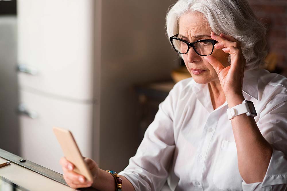 Eine ältere Frau mit Sehschwäche schaut aus weiter Entfernung auf Ihr Smartphone und richtet dabei ihre Brille