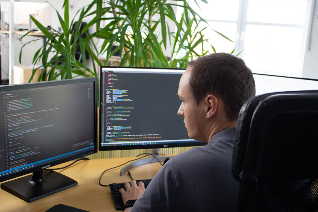 Ein mann sitzt am Schreibtisch an 2 Monitoren auf denen Quellcode zu erkennen ist