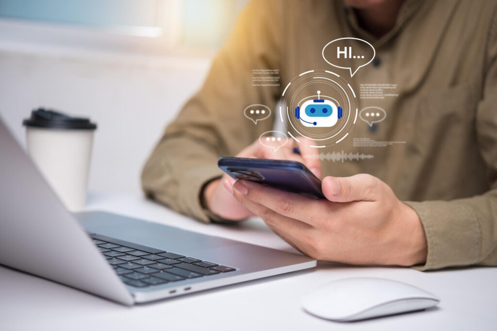 Eine Person sitzt vor einem Laptop an einem Tisch. Sie hält ein Smartphone in der Hand. Über dem Smartphone sind Grafiken die auf einen AI Bot hinweisen.